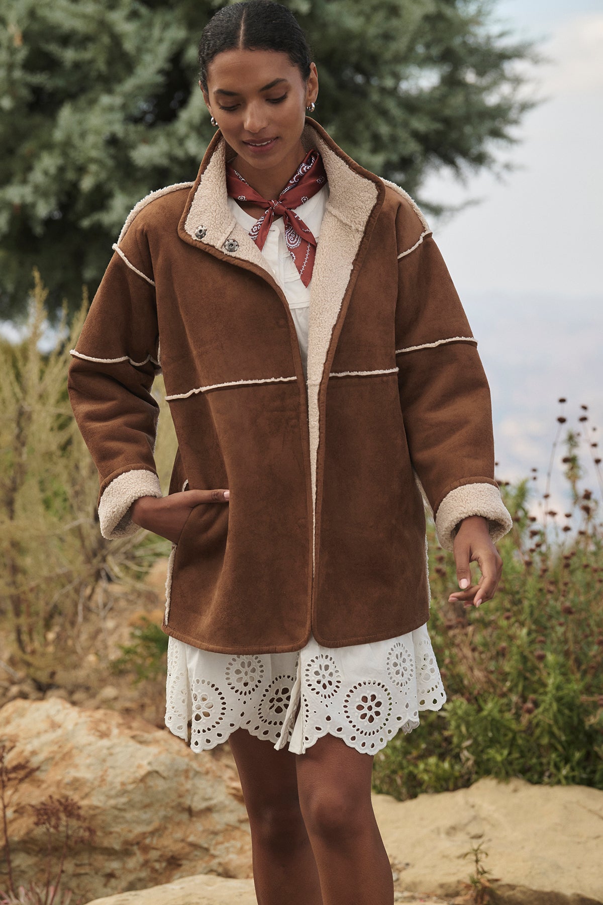   A person wearing the ALBANY LUXE SHERPA REVERSIBLE JACKET by Velvet by Graham & Spencer over a white dress and adorned with a red bandana stands outdoors amid shrubs and rocks, highlighting the eco-friendly faux suede lining that perfectly complements the natural surroundings. 