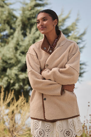 A person outdoors, surrounded by trees, is dressed in the ALBANY LUXE SHERPA REVERSIBLE JACKET from Velvet by Graham & Spencer, showcasing its eco-friendly faux sherpa lining.