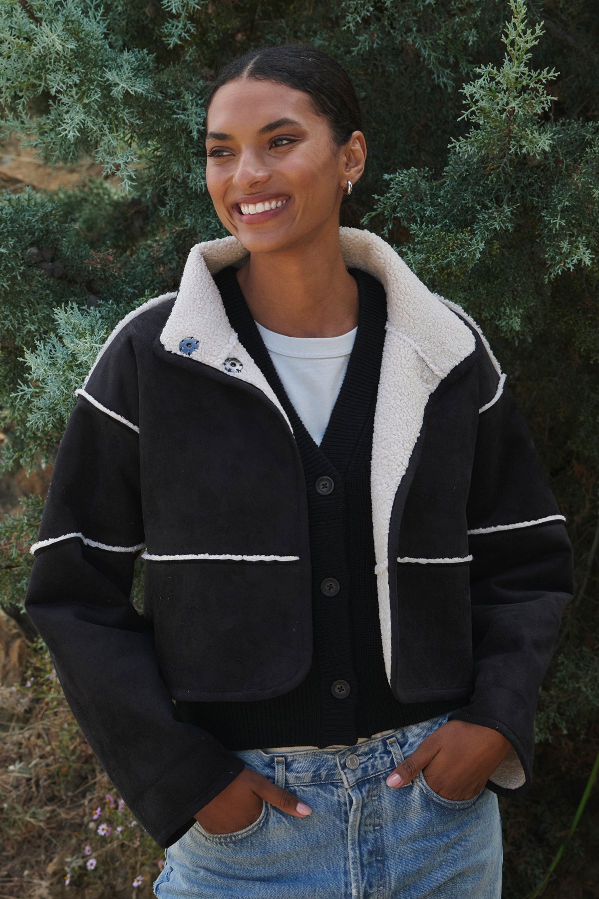   A person is smiling outdoors, enjoying the day in a sleek black sweater and jeans, wearing the KELLY LUXE SHERPA REVERSIBLE JACKET from Velvet by Graham & Spencer. This chic jacket features elegant faux suede detailing that beautifully complements the lush green foliage in the background. 