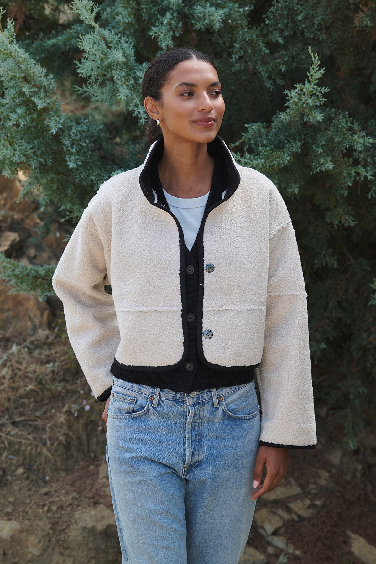   A woman in a stylish KELLY LUXE SHERPA REVERSIBLE JACKET by Velvet by Graham & Spencer and jeans stands outdoors against green foliage, highlighting the versatility of her outfit. 