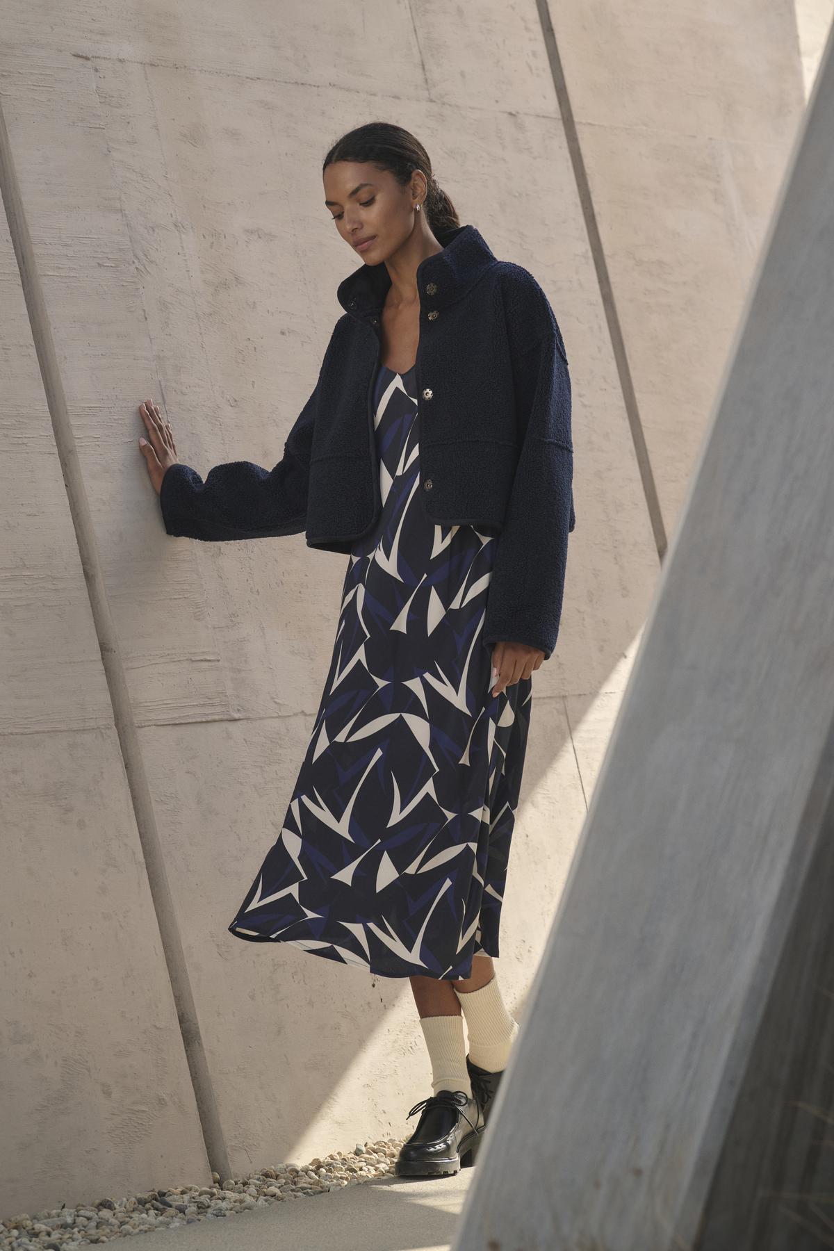   A woman in the PERRY DRESS by Velvet by Graham & Spencer, featuring adjustable straps and a v-neckline, leans against a concrete wall. 