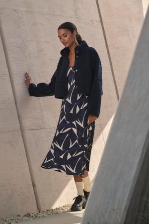 A woman in the PERRY DRESS by Velvet by Graham & Spencer, featuring adjustable straps and a v-neckline, leans against a concrete wall.