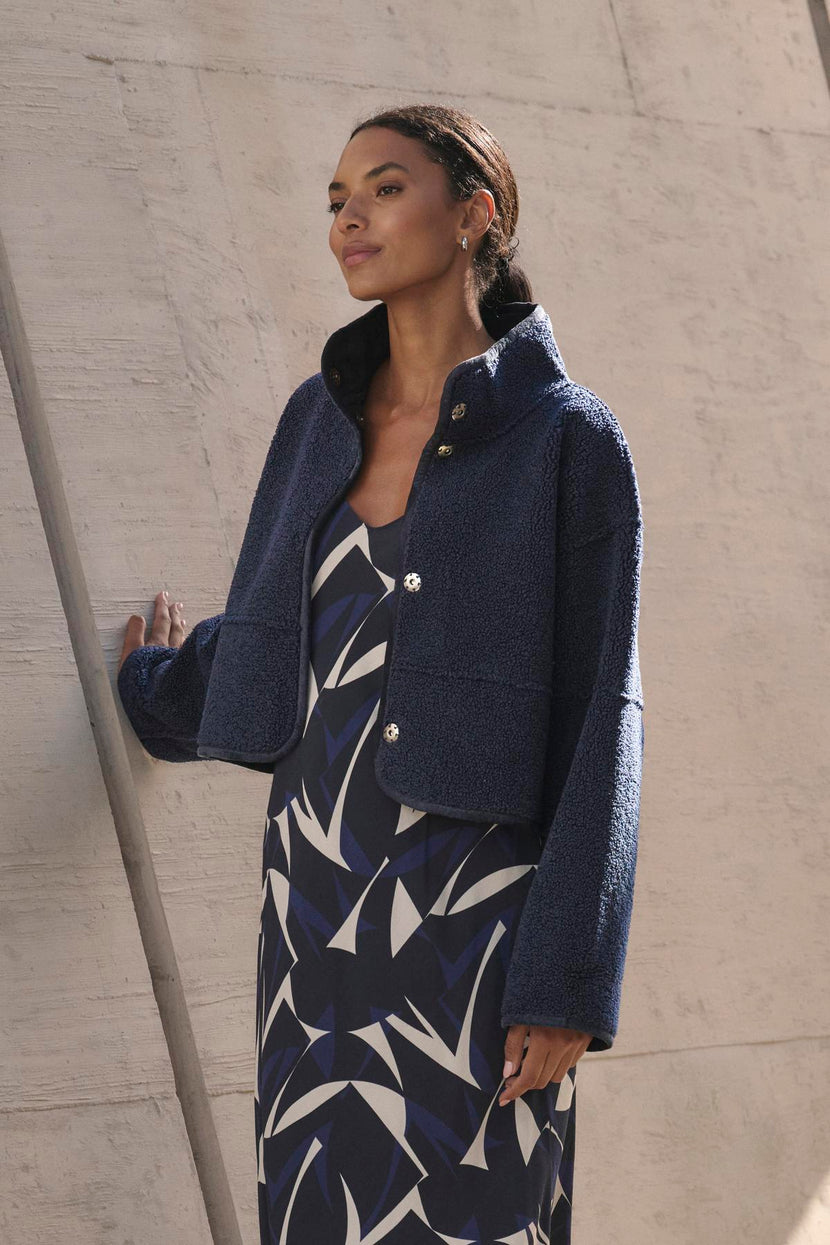 A woman in a patterned dress and the Kelly Luxe Sherpa Reversible Jacket by Velvet by Graham & Spencer stands against a concrete wall, looking upward.