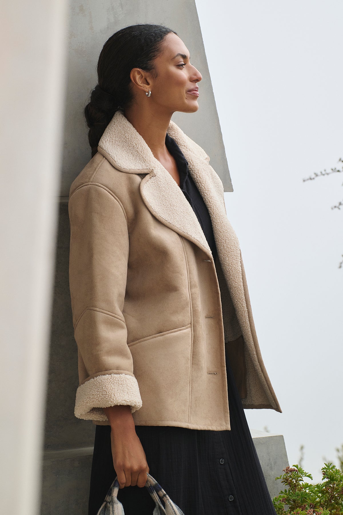   A person wearing the MOLLY LUXE SHERPA JACKET from Velvet by Graham & Spencer over a black outfit, clutching a patterned bag, stands outdoors near a concrete structure, showcasing the timeless appeal of faux suede. 