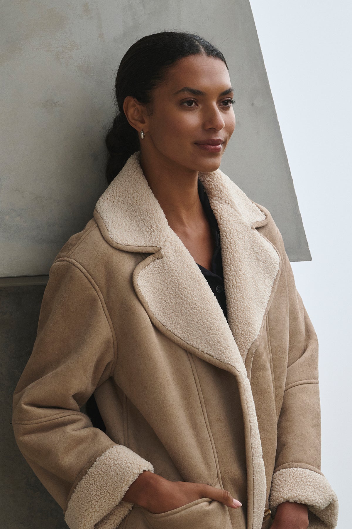   A woman wearing the MOLLY LUXE SHERPA JACKET by Velvet by Graham & Spencer leans against a gray concrete structure, gazing to the side. 