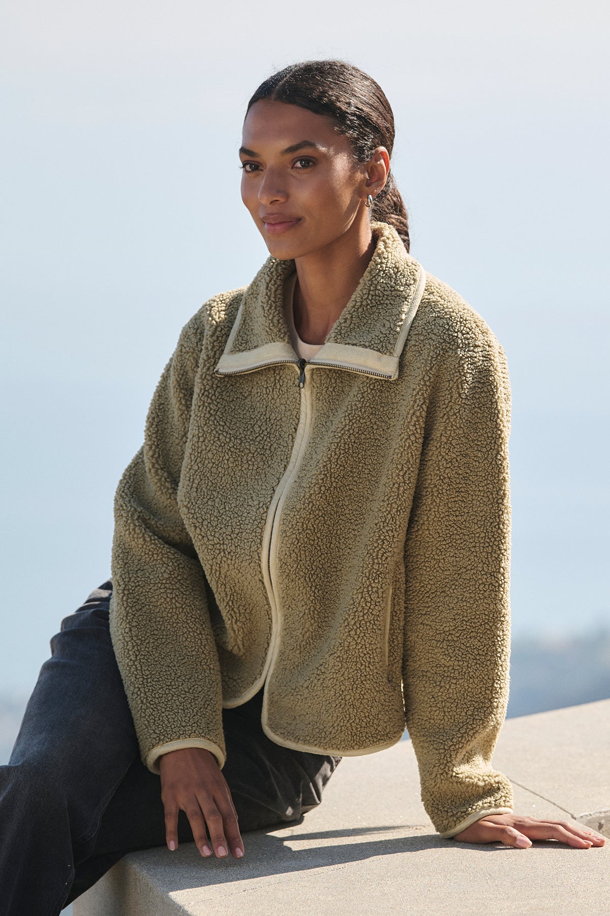   A person sits outdoors in a versatile layering piece—a REBA REVERSIBLE SHERPA JACKET by Velvet by Graham & Spencer paired with dark pants—gazing into the distance. 