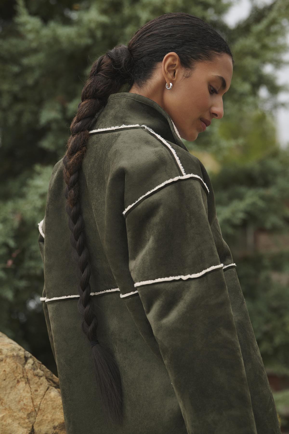   An individual with a lengthy braided ponytail is outdoors, glancing down while wearing the Velvet by Graham & Spencer's eco-friendly ALBANY LUXE SHERPA REVERSIBLE JACKET in olive green. 