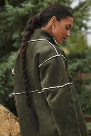 An individual with a lengthy braided ponytail is outdoors, glancing down while wearing the Velvet by Graham & Spencer's eco-friendly ALBANY LUXE SHERPA REVERSIBLE JACKET in olive green.