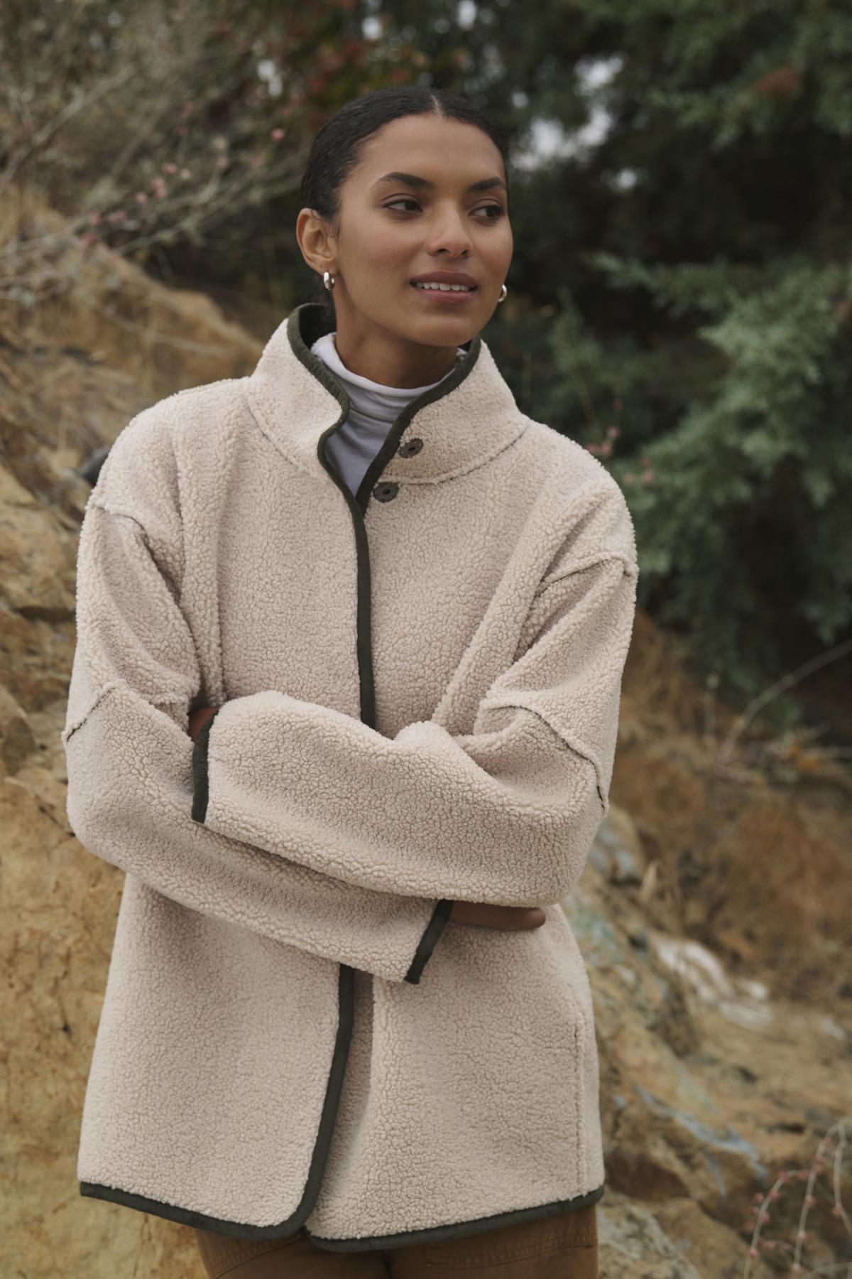 Wearing the ALBANY LUXE SHERPA REVERSIBLE JACKET by Velvet by Graham & Spencer, a person stands outdoors with arms crossed, surrounded by rocky terrain and greenery.-38219215143105