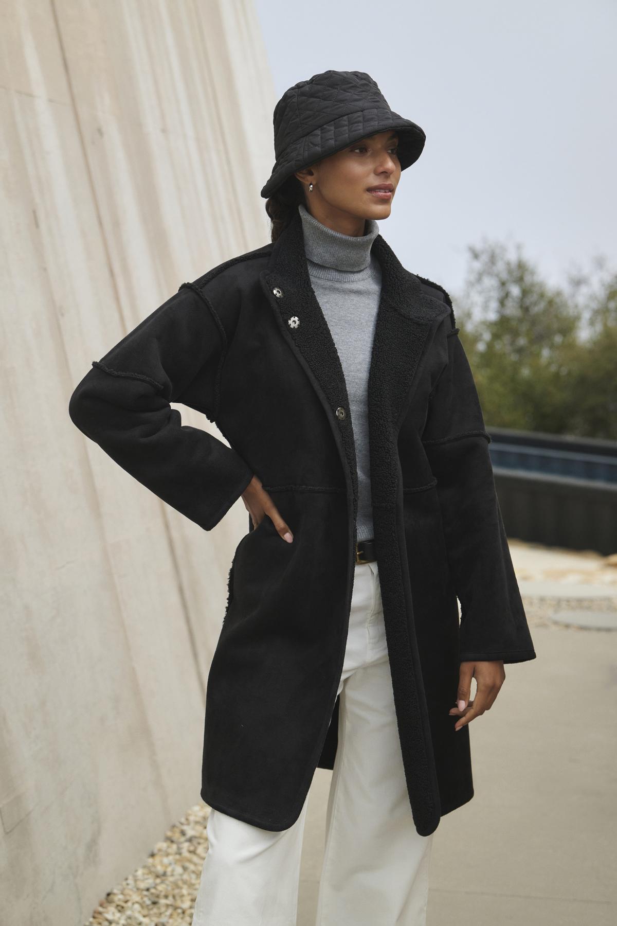   A person wearing a black coat, gray turtleneck, white pants, and black bucket hat stands in an outdoor setting, the ensemble subtly energized by Velvet by Graham & Spencer's Cara Luxe Sherpa Reversible Jacket's hint of faux sherpa detailing. 