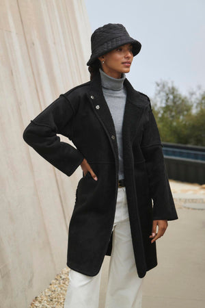 A person wearing a black coat, gray turtleneck, white pants, and black bucket hat stands in an outdoor setting, the ensemble subtly energized by Velvet by Graham & Spencer's Cara Luxe Sherpa Reversible Jacket's hint of faux sherpa detailing.