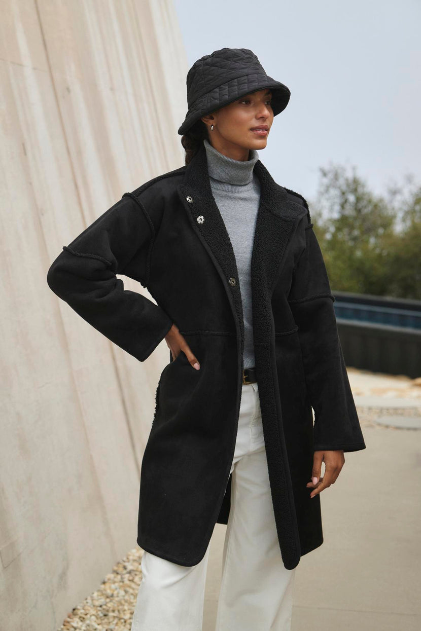 A person wearing a black coat, gray turtleneck, white pants, and black bucket hat stands in an outdoor setting, the ensemble subtly energized by Velvet by Graham & Spencer's Cara Luxe Sherpa Reversible Jacket's hint of faux sherpa detailing.