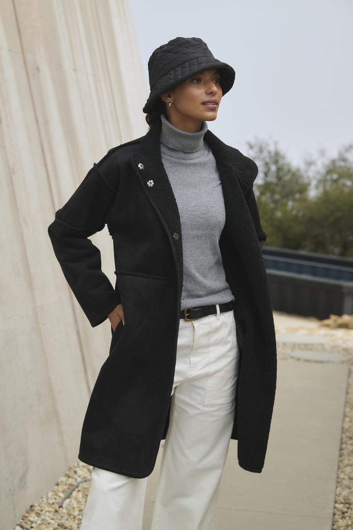   A person wearing a black coat, the KORIE TURTLENECK SWEATER from Velvet by Graham & Spencer, white pants, and a black hat stands outdoors near a building with white walls and a stone pathway. 