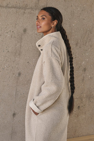 Individual with long braided hair wearing a light-colored, textured Cara Luxe Sherpa Reversible Jacket by Velvet by Graham & Spencer, standing against a concrete wall.