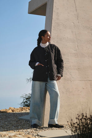 A person in a PAITYN QUILTED JACKET by Velvet by Graham & Spencer and jeans stands outdoors near a modern concrete structure, with a clear sky and rocky ground in the background.