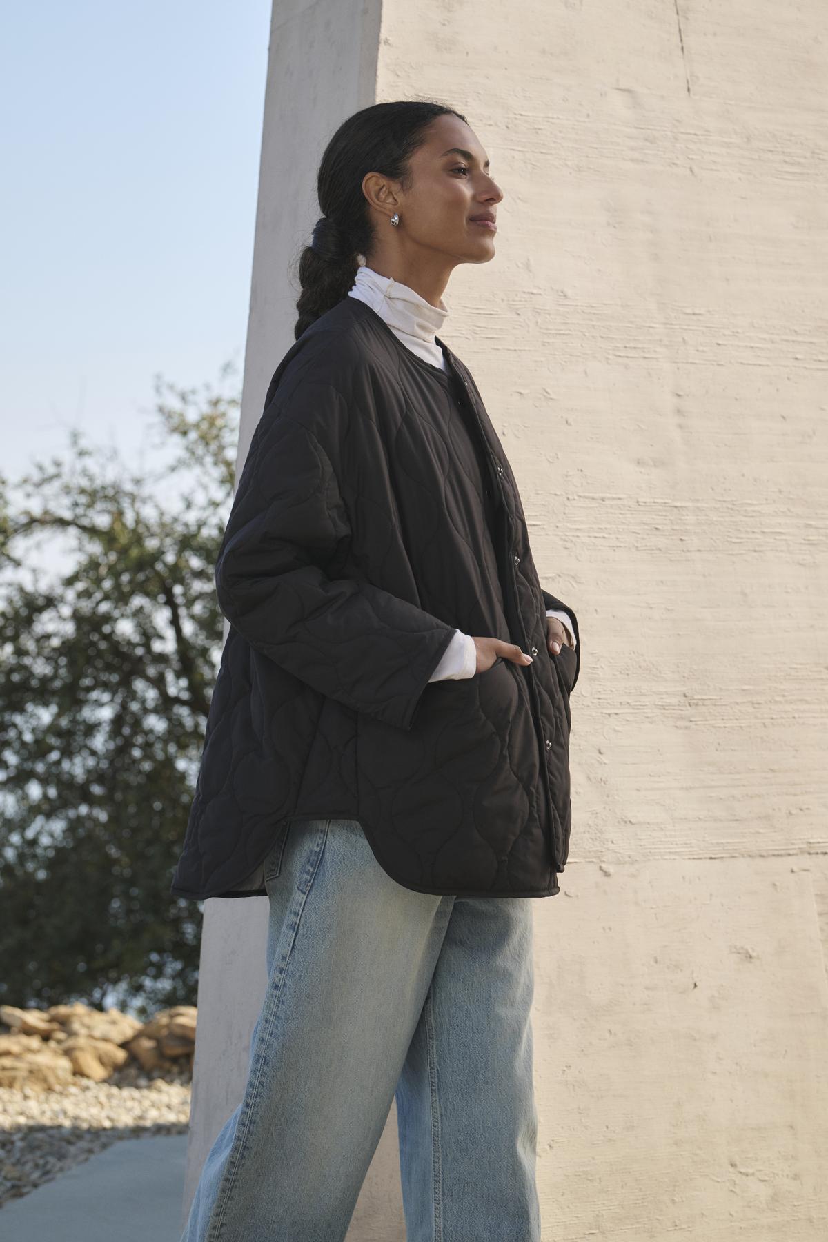   A woman dressed in the PAITYN QUILTED JACKET by Velvet by Graham & Spencer, paired with light jeans, stands in profile with her hands in her pockets against a light wall outside. 