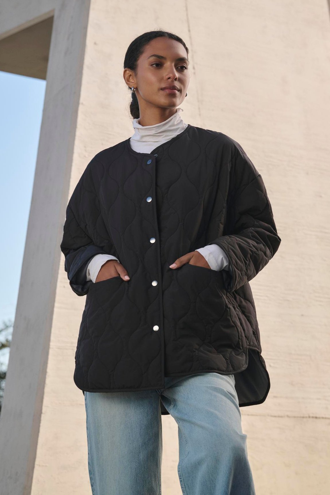   A person wearing the PAITYN QUILTED JACKET from Velvet by Graham & Spencer and jeans stands outdoors near a large concrete structure, looking away from the camera. 