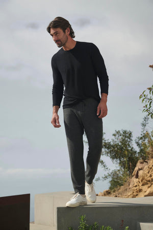 A man in a black long-sleeve shirt and the ZEALAND SWEATPANT by Velvet by Graham & Spencer strolls outdoors on a concrete path, with greenery and sky creating a serene backdrop.