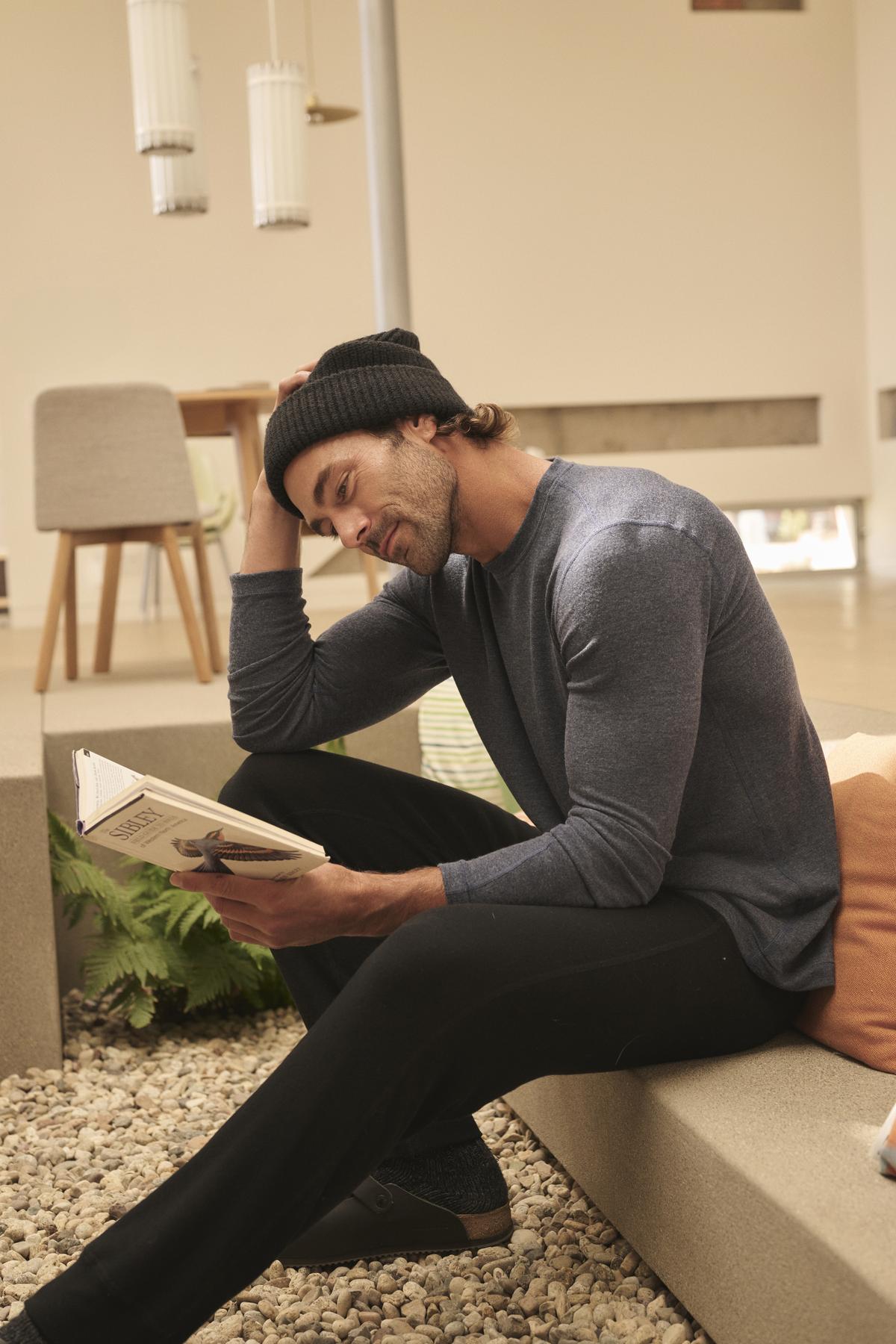   A person wearing a beanie, long-sleeve shirt, and ZEALAND SWEATPANT by Velvet by Graham & Spencer sits on a bench reading a book in a modern, light-filled room. 