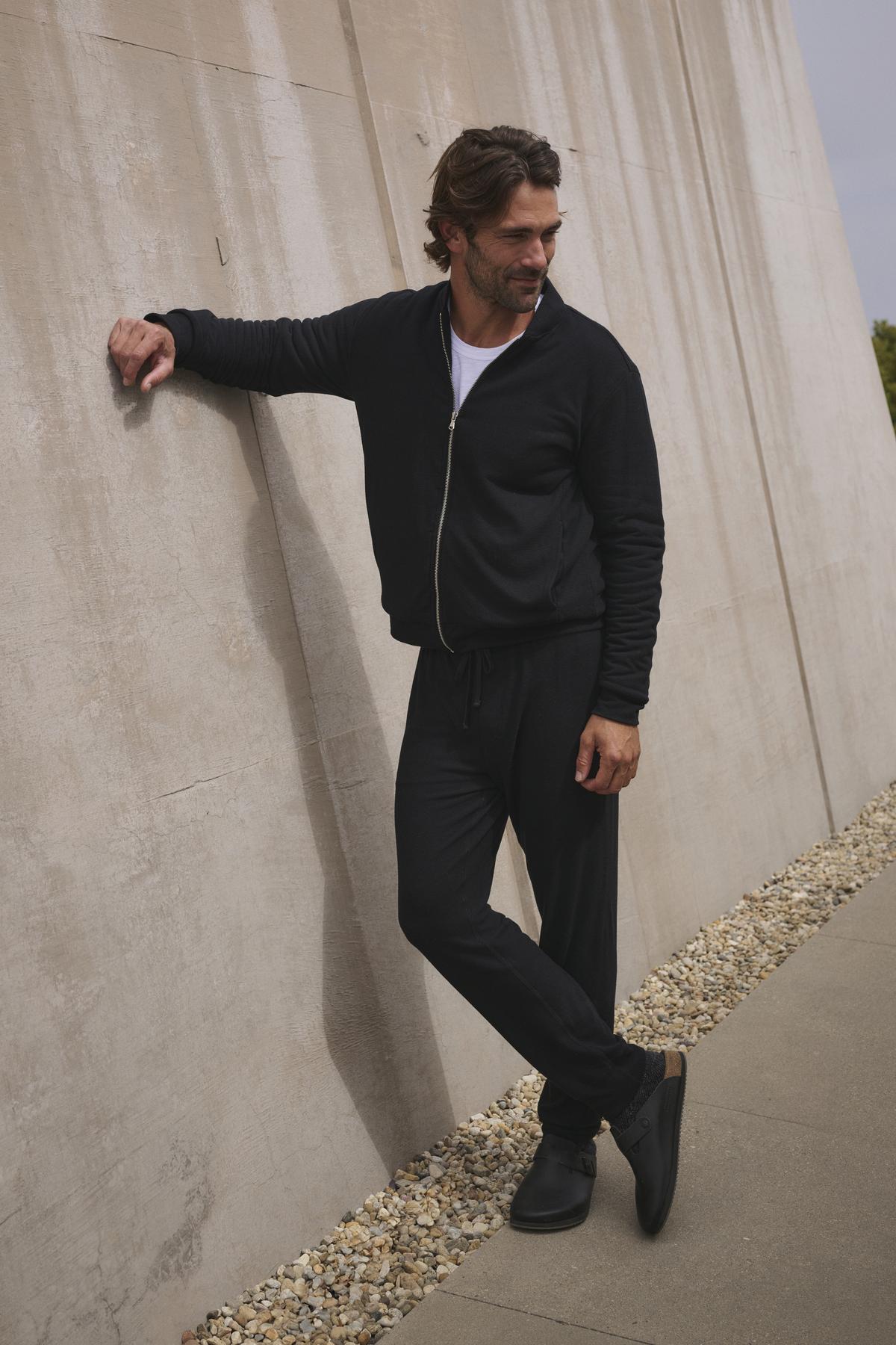   Dressed in a black ensemble featuring the ZEALAND SWEATPANT by Velvet by Graham & Spencer, he leans casually with his right hand against a concrete wall while standing on a pebble-lined path. 