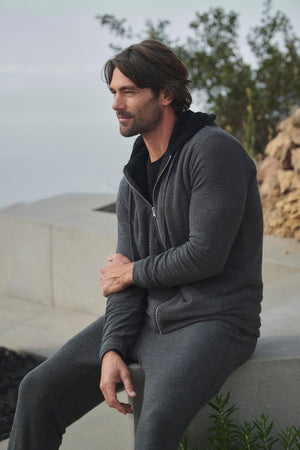 A man dressed in ZEALAND SWEATPANT by Velvet by Graham & Spencer sits outdoors on a concrete ledge with greenery in the background, wearing a gray hoodie and glancing to the side.