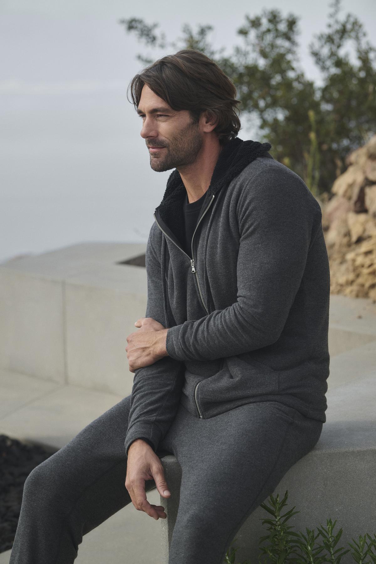 A man wearing the SALVADORE HOODIE by Velvet by Graham & Spencer, designed with ultra-soft cozy jersey fabric, sits on a stone ledge outdoors with a neutral expression, framed by plants in the background.-38282327654593