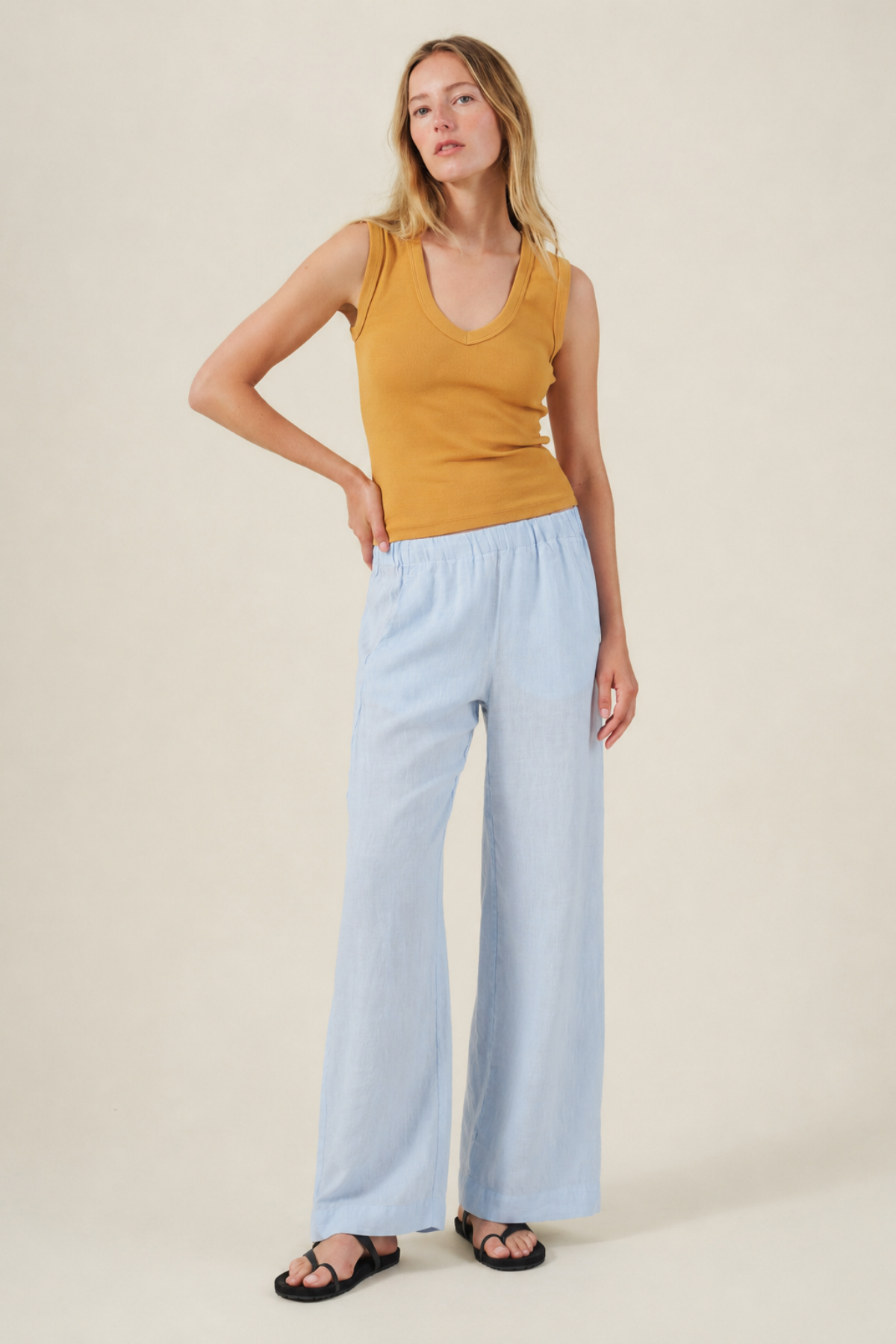 A woman stands against a light background wearing a mustard yellow sleeveless top, Velvet by Graham & Spencer's STEPHANIE LINEN PANT with a wide leg and elastic waist, and black sandals.-41681795219649