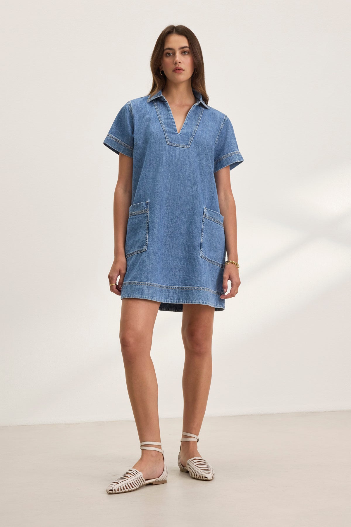 A woman models the DELANA DENIM DRESS by Velvet by Graham & Spencer, featuring short sleeves and two large pockets. She pairs it with white sandals and a bracelet, all set against a plain white background.-38802060411073