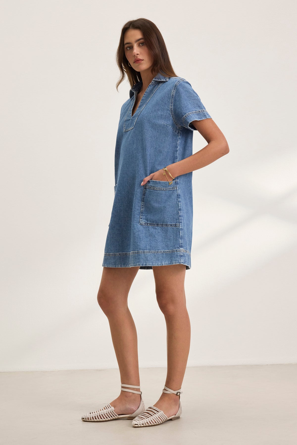 A person wearing the DELANA DENIM DRESS by Velvet by Graham & Spencer, featuring short sleeves and front pockets, paired with white strappy sandals, stands sideways against a plain background.-38802060345537