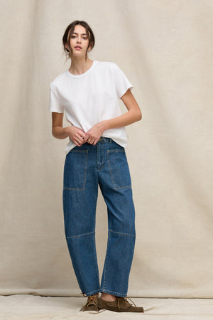 A woman stands against a beige background wearing the FARAH DENIM PANT by Velvet by Graham & Spencer, featuring high-waisted, cropped, curved-leg denim with front patch pockets, paired with a plain white t-shirt and brown lace-up shoes.