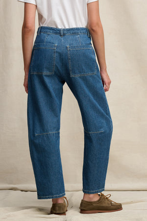 A person stands with their back to the camera wearing Velvet by Graham & Spencer’s FARAH DENIM PANT—loose-fitting blue jeans with large patch pockets and curved legs—paired with a white t-shirt and brown shoes against a neutral background.