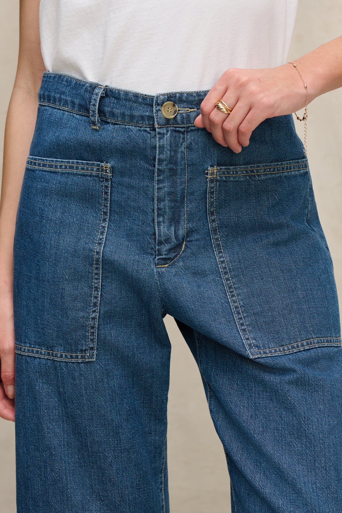   A person wears the FARAH DENIM PANT by Velvet by Graham & Spencer—cropped denim pants with large front patch pockets—paired with a white shirt, holding the waistband with one hand. 