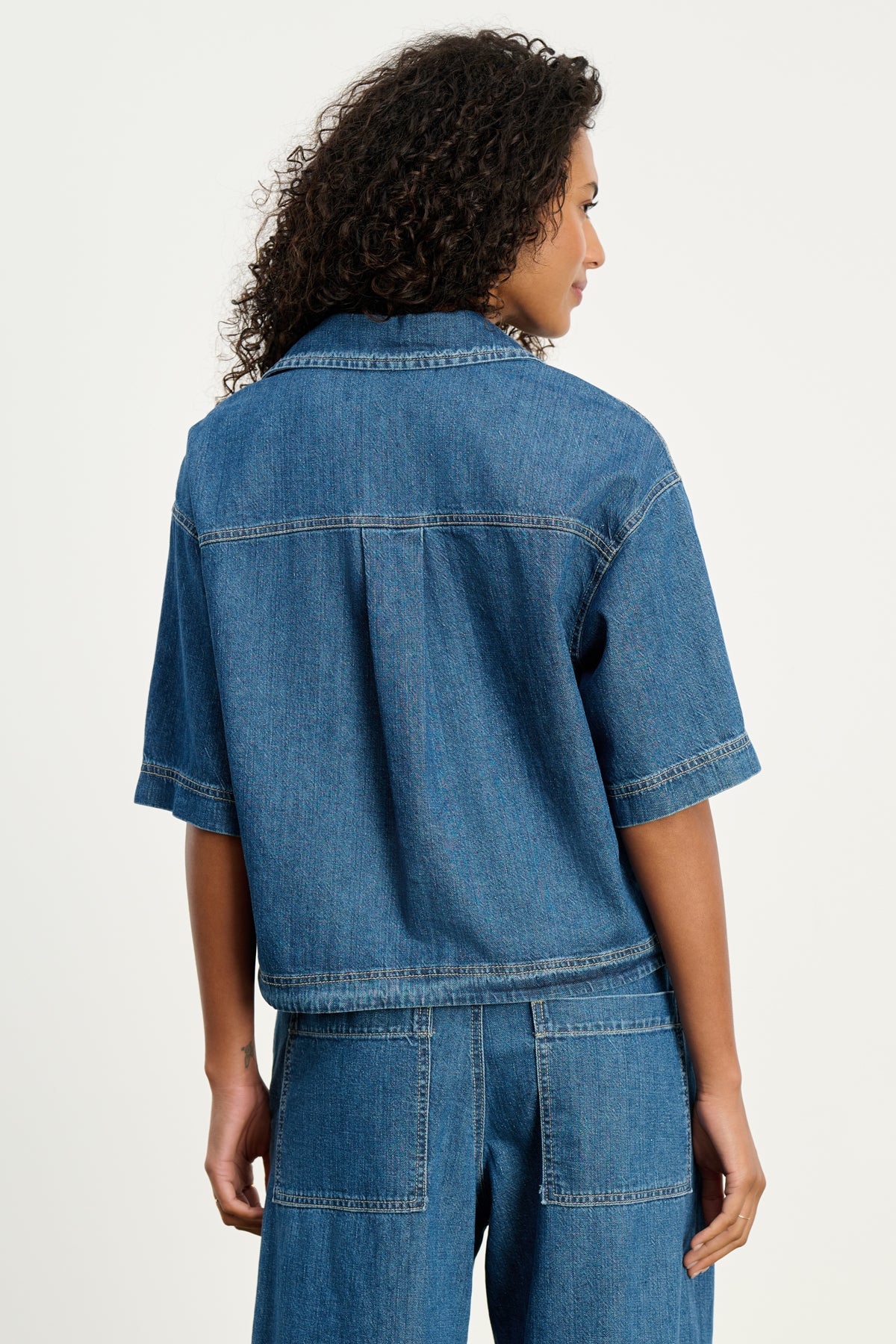 A person with curly hair models the KADDY DENIM TOP by Velvet by Graham & Spencer and matching relaxed-fit denim pants, shown from the back against a plain light background.-39800343724225