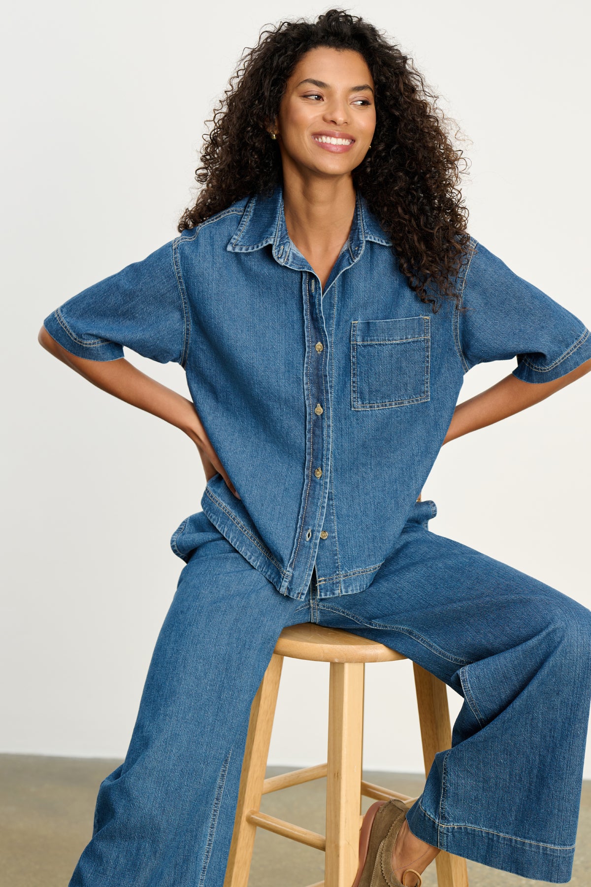   A woman with curly hair sits on a wooden stool, wearing the Velvet by Graham & Spencer KADDY DENIM TOP with a chest pocket and matching wide-leg denim pants. 