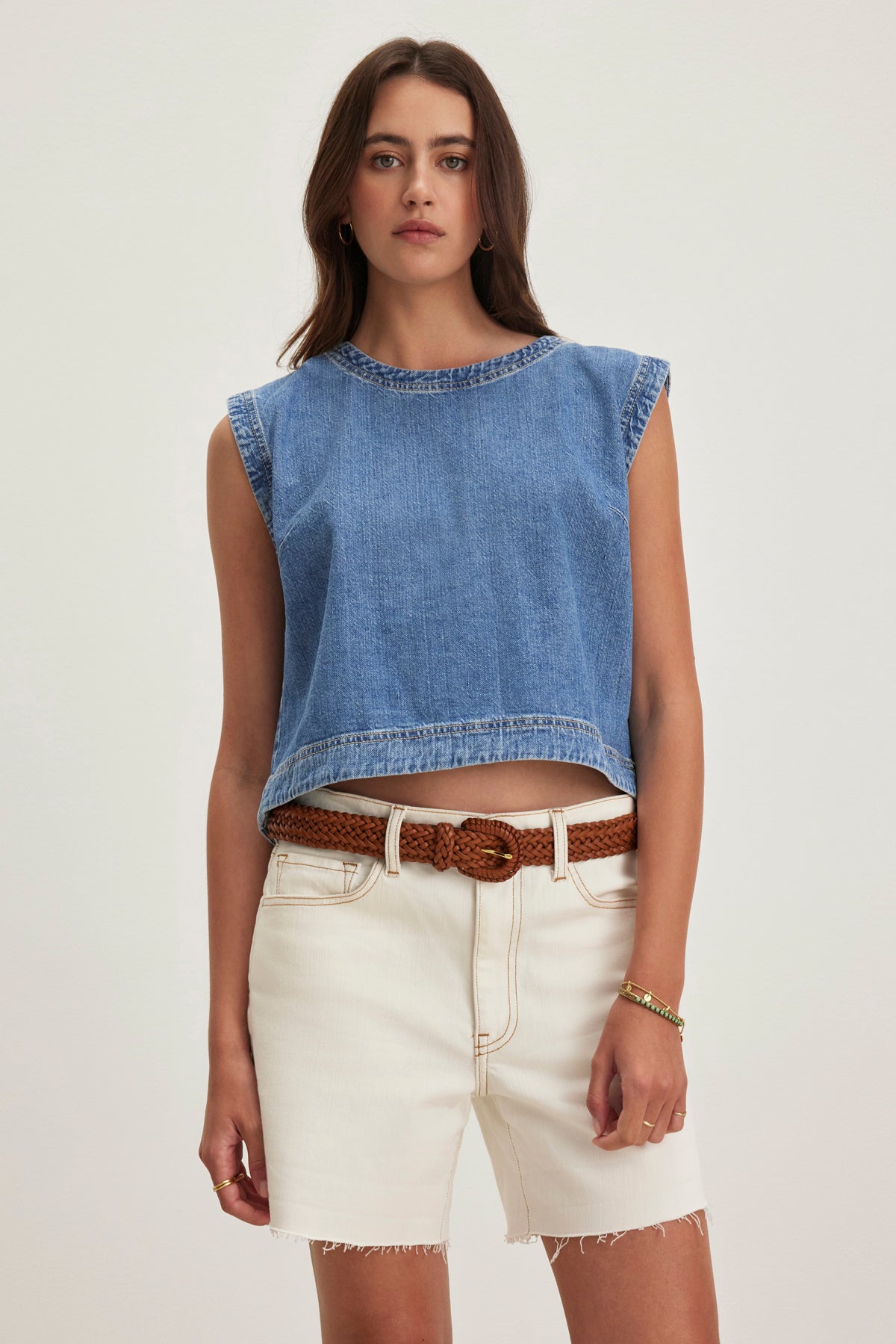 A person wearing the sleeveless LOREN DENIM TOP by Velvet by Graham & Spencer, cream-colored shorts, and a brown braided belt stands against a plain background.-38820684660929