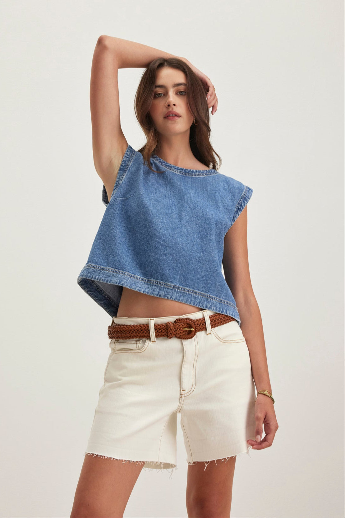 A woman poses in the LOREN DENIM TOP by Velvet by Graham & Spencer, paired with white frayed shorts and a brown braided belt. With her left arm raised, she stands against a plain background.-38820684333249
