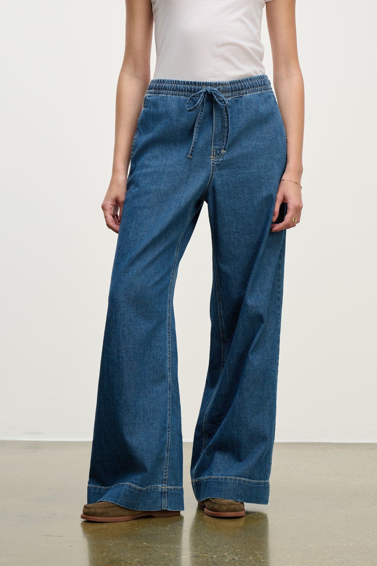 A model wears the MIRA DENIM PANT by Velvet by Graham & Spencer—wide-leg denim pants with a drawstring waist—paired with a white sleeveless top, standing on a light brown floor against a plain white background.-39742338990273