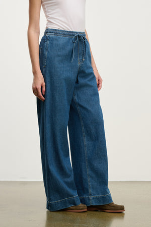 A person stands on a speckled floor against a plain background, wearing the MIRA DENIM PANT by Velvet by Graham & Spencer with a white sleeveless top and brown shoes.