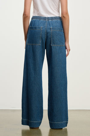 Model wears the MIRA DENIM PANT by Velvet by Graham & Spencer—a wide-leg denim pant with a drawstring waist and large back pockets—styled with a white shirt, photographed from behind against a plain background.