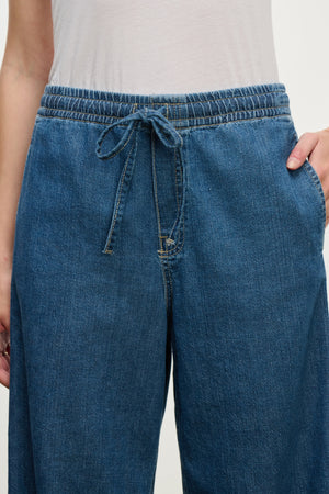 Model wears the MIRA DENIM PANT by Velvet by Graham & Spencer—blue wide-leg pants with a drawstring and elastic waistband, paired with a white top. Shown waist to upper thigh, one hand in pocket.
