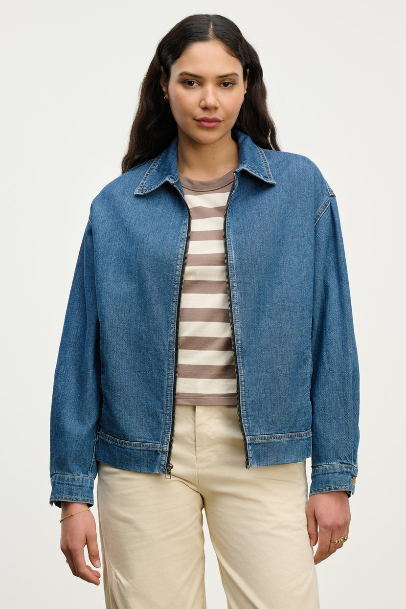 A woman with long dark hair models the PORTER DENIM ZIP-UP JACKET by Velvet by Graham & Spencer over a beige and white striped shirt and light pants, standing against a plain background.