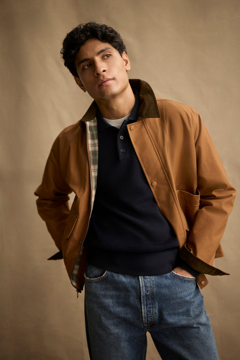 A man stands with his hands in his pockets against a plain beige background, wearing the DESREN JACKET by Velvet by Graham & Spencer—a brown cotton canvas jacket with a corduroy collar—over a dark sweater and blue jeans.