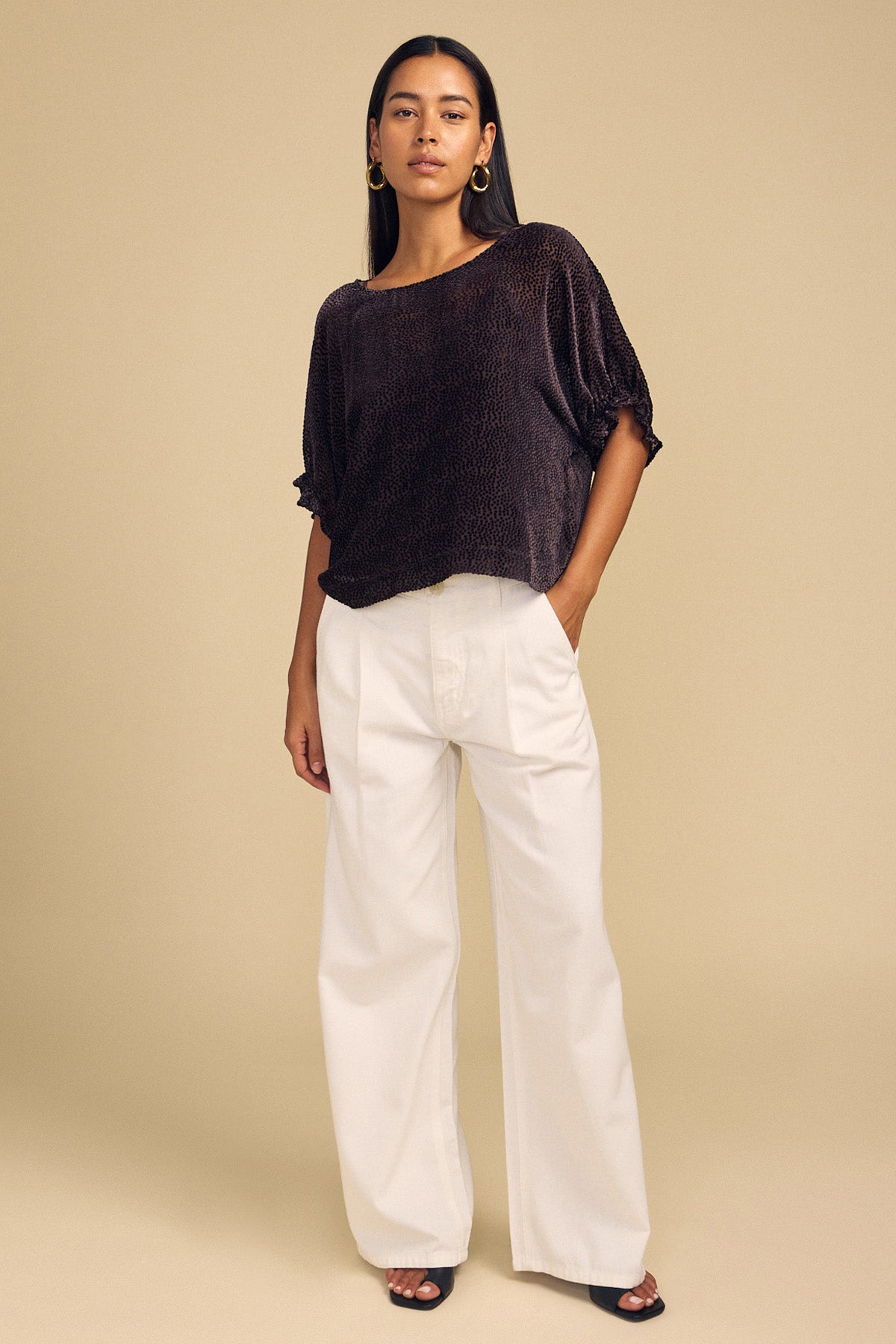 A woman wearing a dark, short-sleeved Velvet by Graham & Spencer ADDIE DEVORE DOT BLOUSE with a subtle dotted texture and wide-leg white trousers.-46488075829441