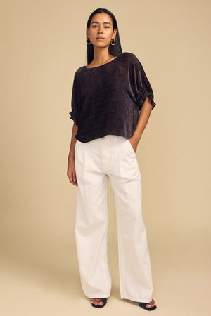 A woman wearing a dark, short-sleeved Velvet by Graham & Spencer ADDIE DEVORE DOT BLOUSE with a subtle dotted texture and wide-leg white trousers.