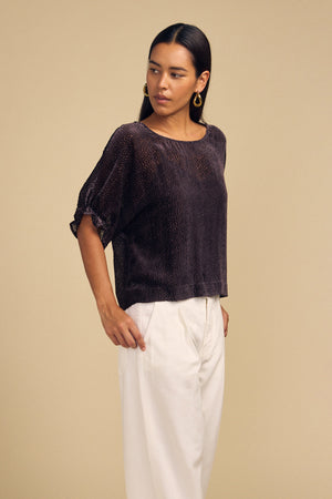 A person wears a Velvet by Graham & Spencer ADDIE DEVORE DOT BLOUSE with short sleeves and a subtle dotted texture, paired with white trousers.