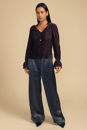 A woman wearing an EMERSON DEVORE DOT BLOUSE in dark silk with a delicate dot pattern, ruffled collar and cuffs, and wide-leg trousers.