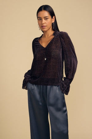 A person wearing a dark brown, long-sleeved Velvet by Graham & Spencer EMERSON DEVORE DOT BLOUSE with a V-neckline and ruffled cuffs, paired with dark gray trousers.