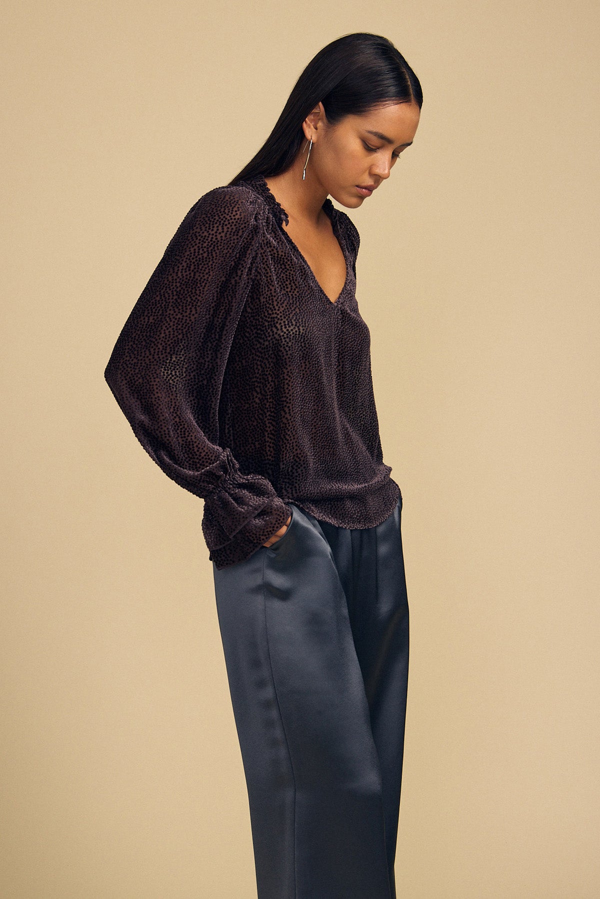 A person wearing an EMERSON DEVORE DOT BLOUSE, a long-sleeved, v-neck silk devoré top with a dark dotted pattern, paired with dark trousers.