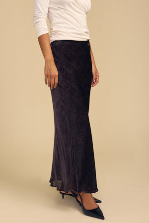 A person wearing a QUINN DEVORE DOT SKIRT, a white long-sleeved top, and black pointed-toe heels.
