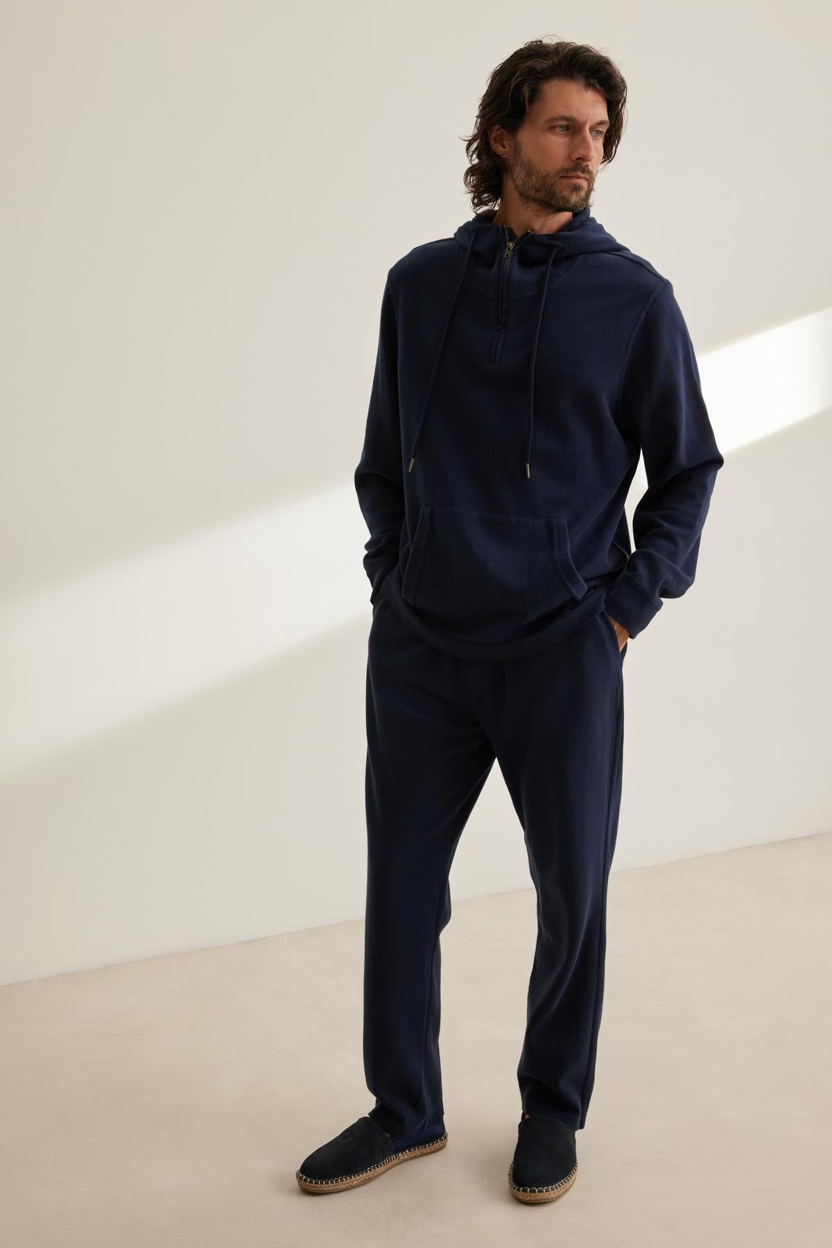   A man in Velvet by Graham & Spencer's navy BLAYNE JOGGER hoodie and matching pants stands relaxed against a plain backdrop. 